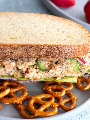 A salmon salad sandwich made with Greek yogurt and healthy ingredients, served on a white plate with a side of pretzels.
