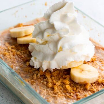 Banana pudding overnight oats in a transparent container, topped with banana slices, crushed graham crackers and whipped cream.