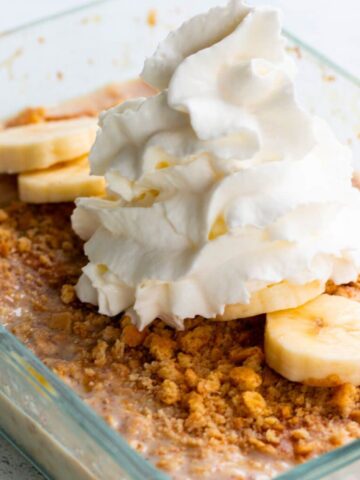 Banana pudding overnight oats in a transparent container, topped with banana slices, crushed graham crackers and whipped cream.