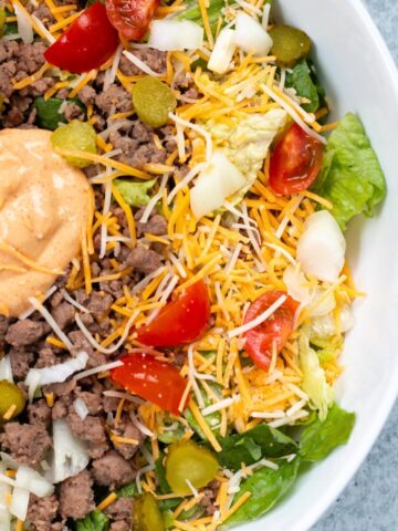 A Big Mac bowl served in a white bowl, featuring ground beef, cheese, pickles, cherry tomatoes, and cheese burger salad dressing.