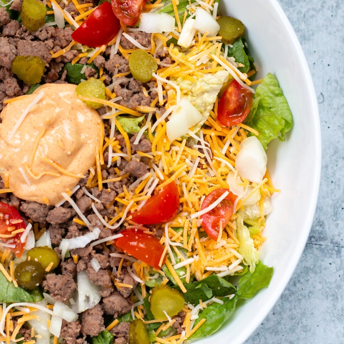 A Big Mac bowl served in a white bowl, featuring ground beef, cheese, pickles, cherry tomatoes, and cheese burger salad dressing.